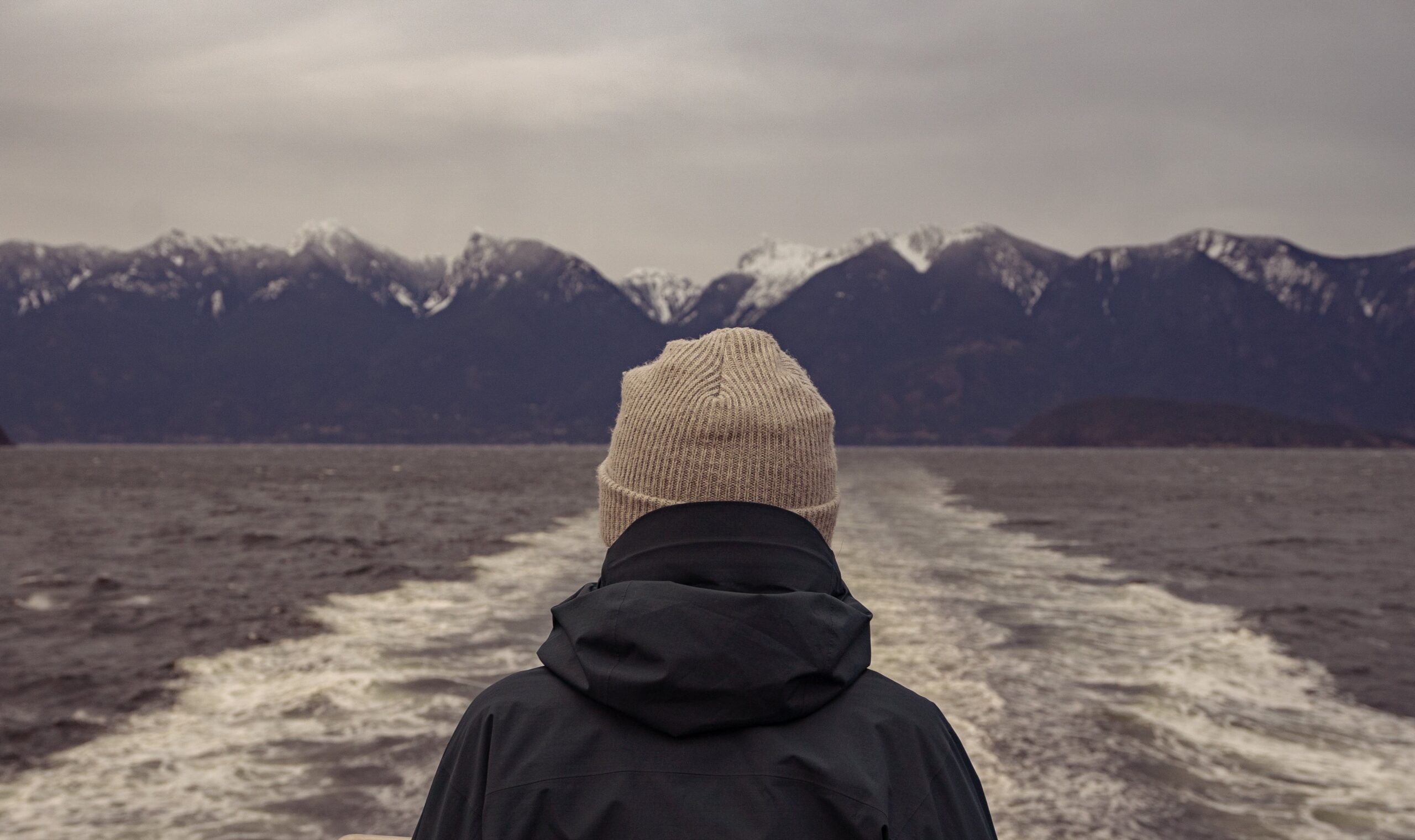 Boater wearing a knit hat and jacket looks across the boat’s wake toward snow-capped mountains.